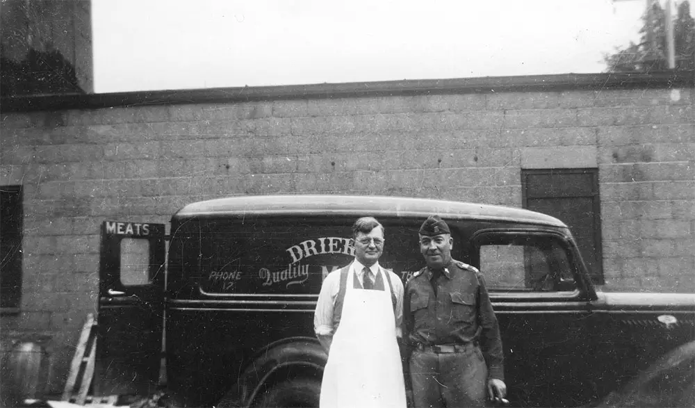 Black-and-white photo of a butcher standing beside the vintage Drier's Meat Market delivery van