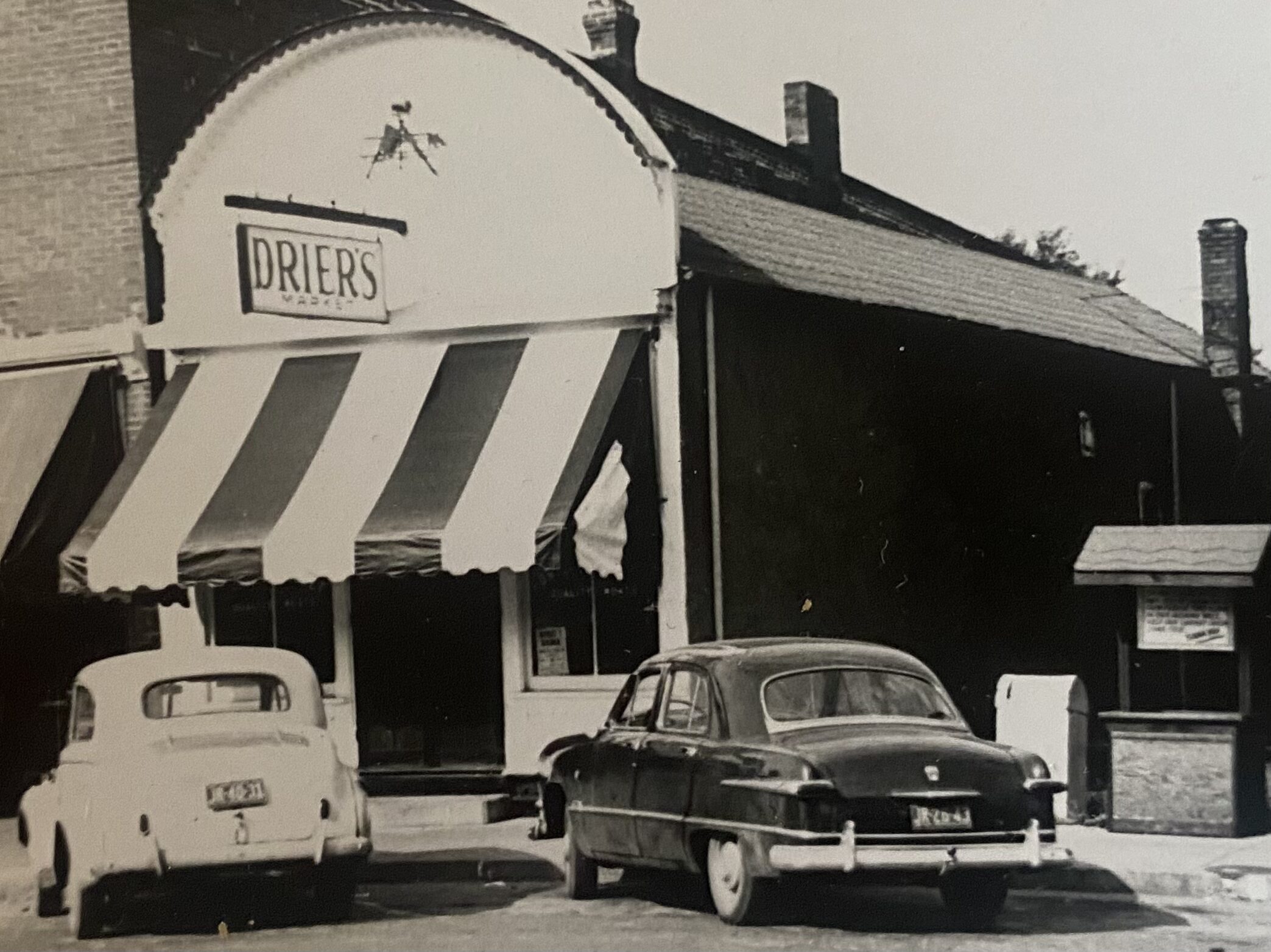 Black and white historic photo of the Drier's Market storefront in Three Oaks, Michigan, with a striped awning and classic cars parked outside