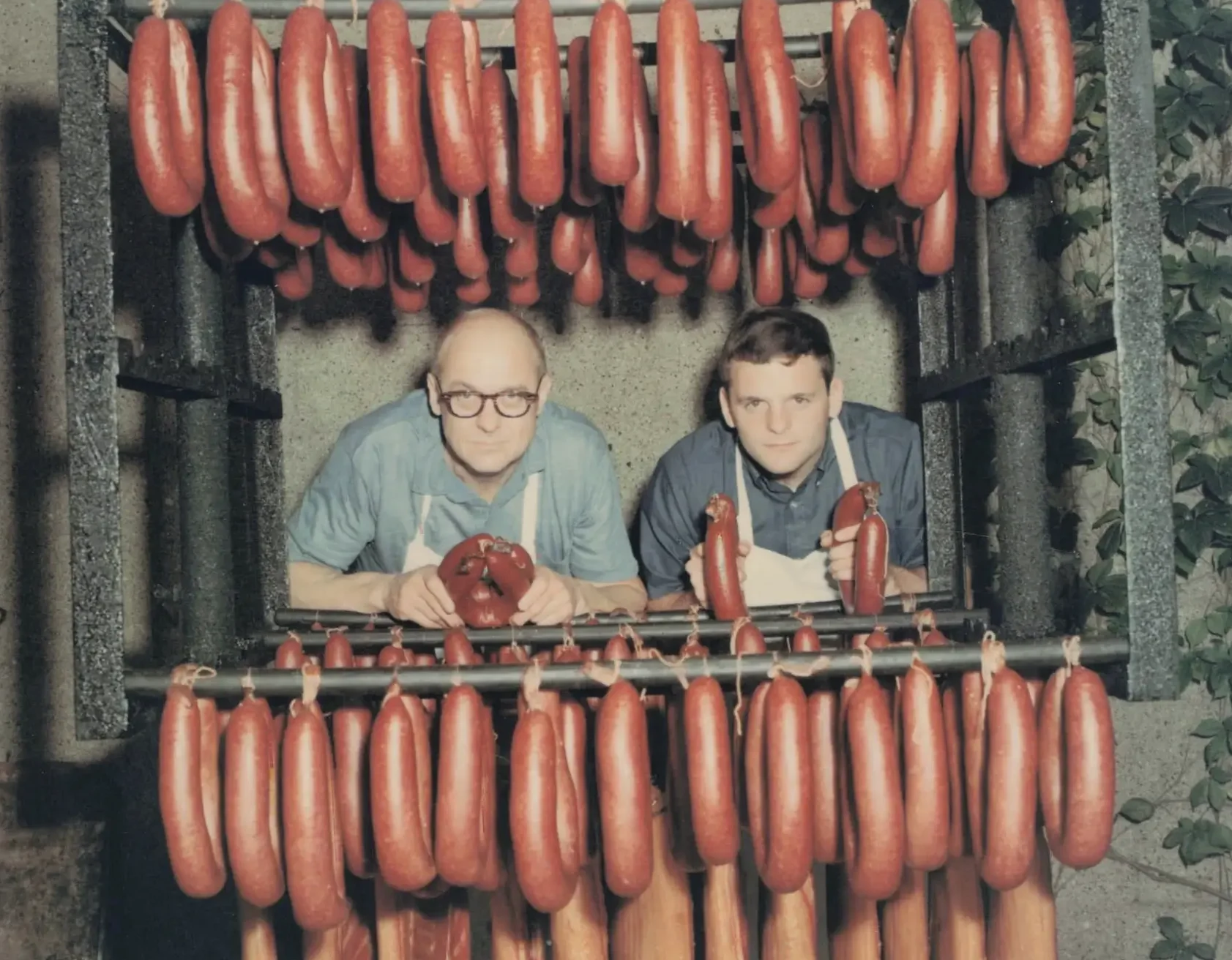 Color photo of an older man and a child at the Drier's Meat Market counter beneath hanging rings of baloney